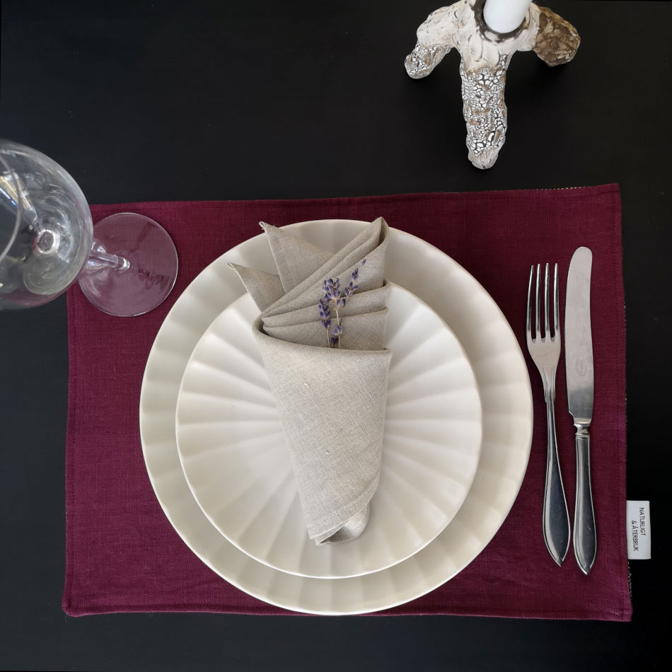 Vinröd bordstablett i linne dukat med porslin och bestick samt naturfärgad servett.