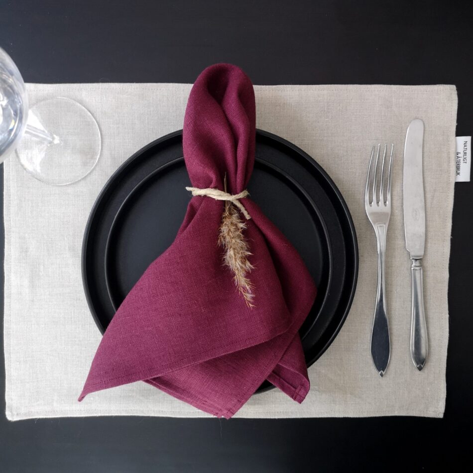 Naturfärgad bordstablett i linne dukat med porslin och bestick samt vinröd servett.