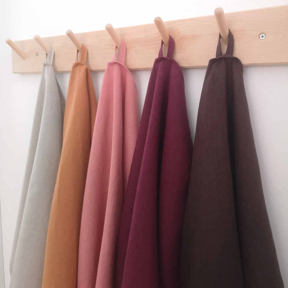 Fem stycken handdukar i linne i olika färger hänger på träkrokar tillsammans med torkad lavendel och en äldre mattpiska.